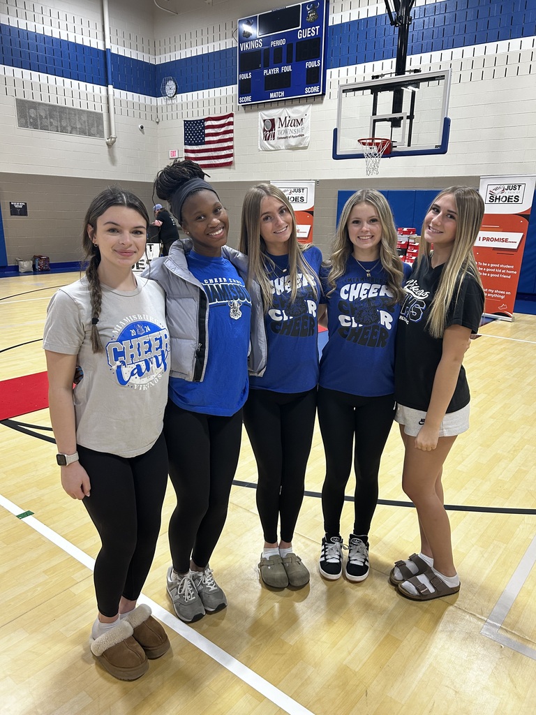 Five women, wearing matching blue shirts, pose together on a basketball court. The background includes a basketball hoop, a red floor, and blue and white walls.