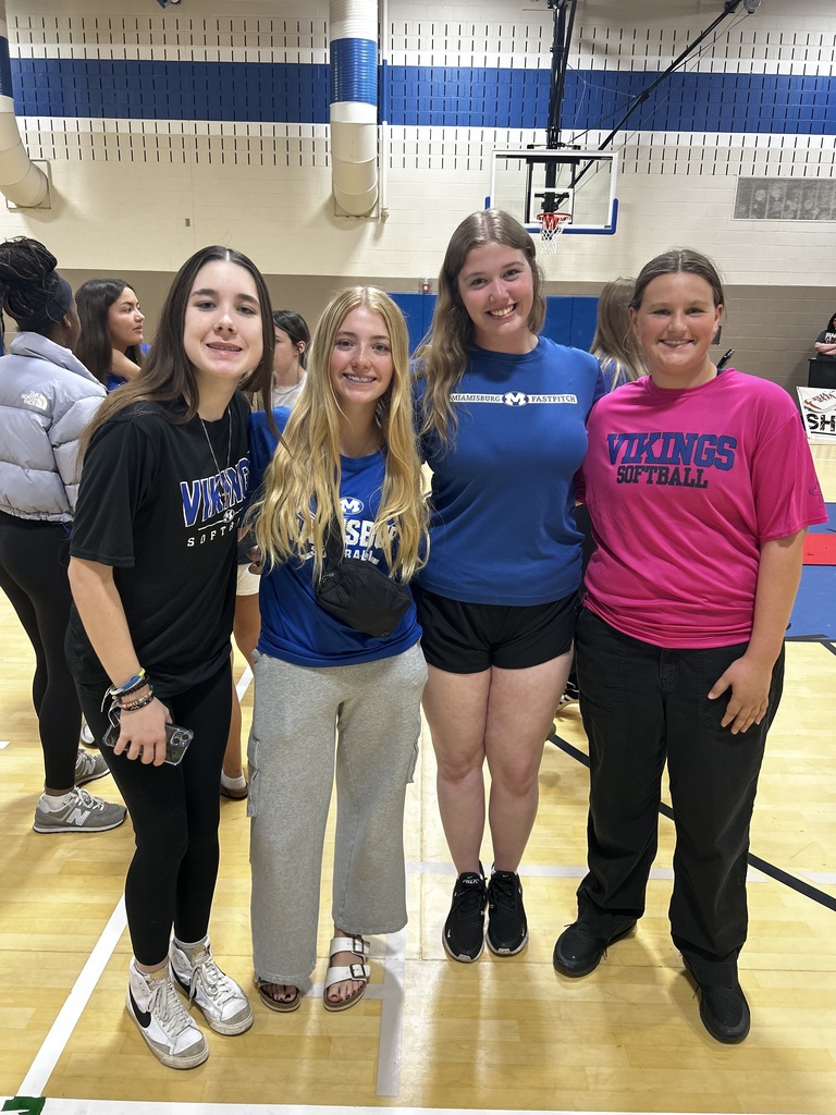Four women in athletic wear stand on a basketball court with a hoop. Two wear black shirts, one blue, and one pink.