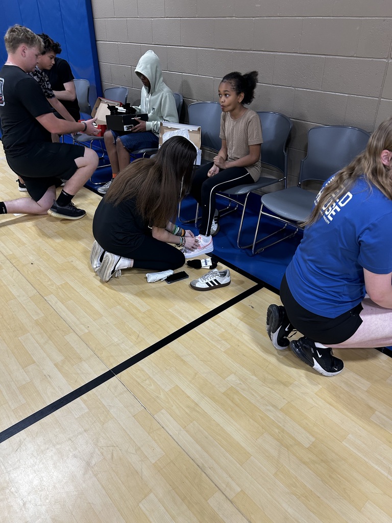 Several people are seated or kneeling on a wooden floor, likely engaged in an activity. One person is examining another's shoes.
