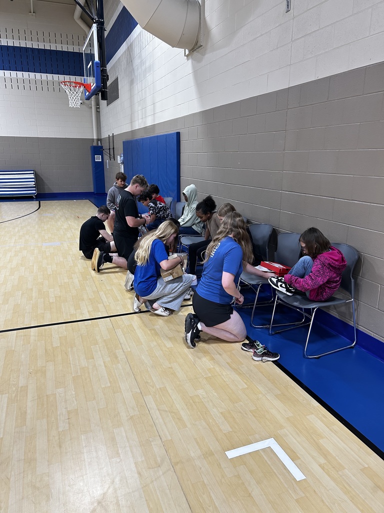 A group of people, including adults and children, are sitting on chairs along the wall of an indoor basketball court.