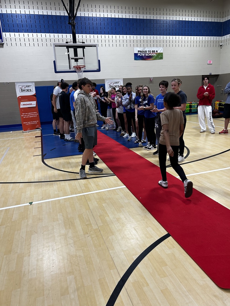 People line up on a red carpet in a gymnasium, with a basketball hoop in the background.