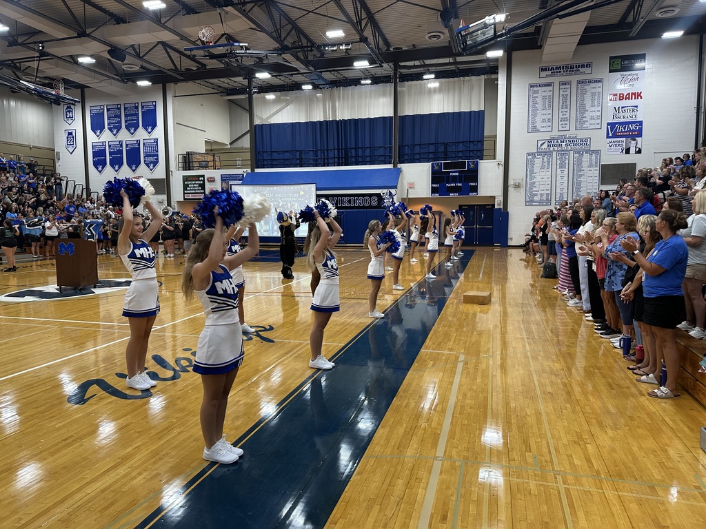 A cheerleading squad performs in a gymnasium. Six cheerleaders are on the blue floor. Audience members line the sidelines.