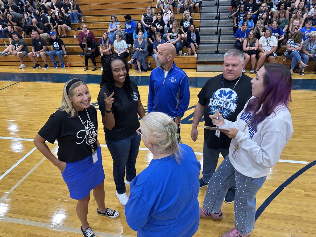 Group of people, including adults and teens, on a basketball court. One person holds a microphone.