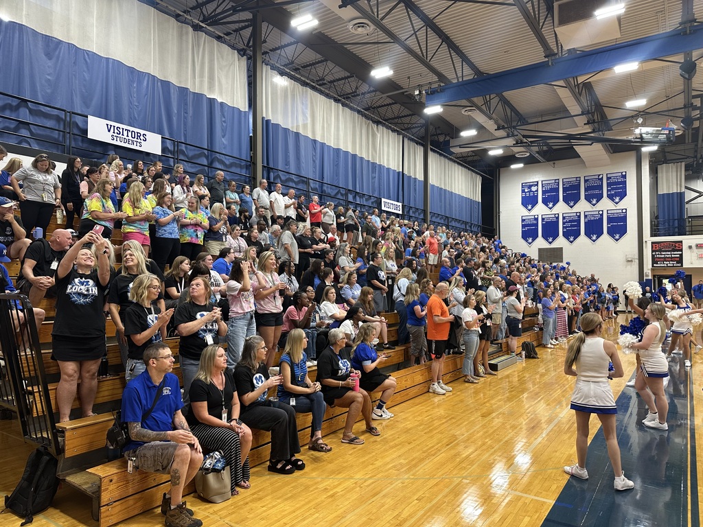 A large crowd of people in a gym, watching cheerleaders on the basketball court. The bleachers are full.