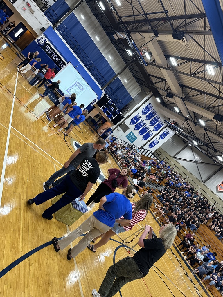 A crowd of people in a gymnasium with a large screen. Some are standing and others are seated.