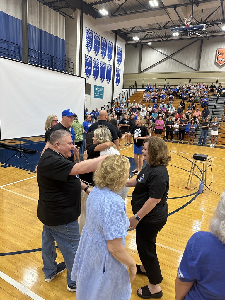 A group of people stand in a gymnasium, some hugging, with a projector screen and bleachers filled with spectators.
