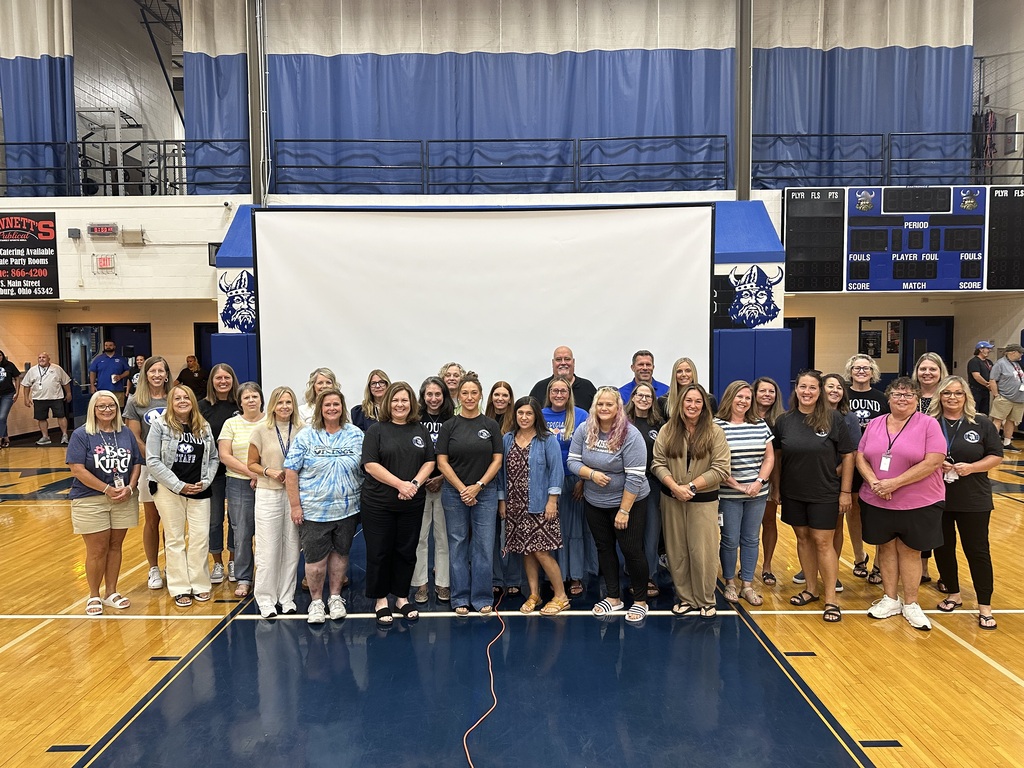 Group of people standing in a row, smiling, in a gymnasium. Blue curtains behind them.