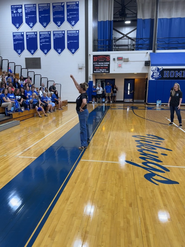 Two people stand on a basketball court, one with arms raised, as an audience sits in bleachers.