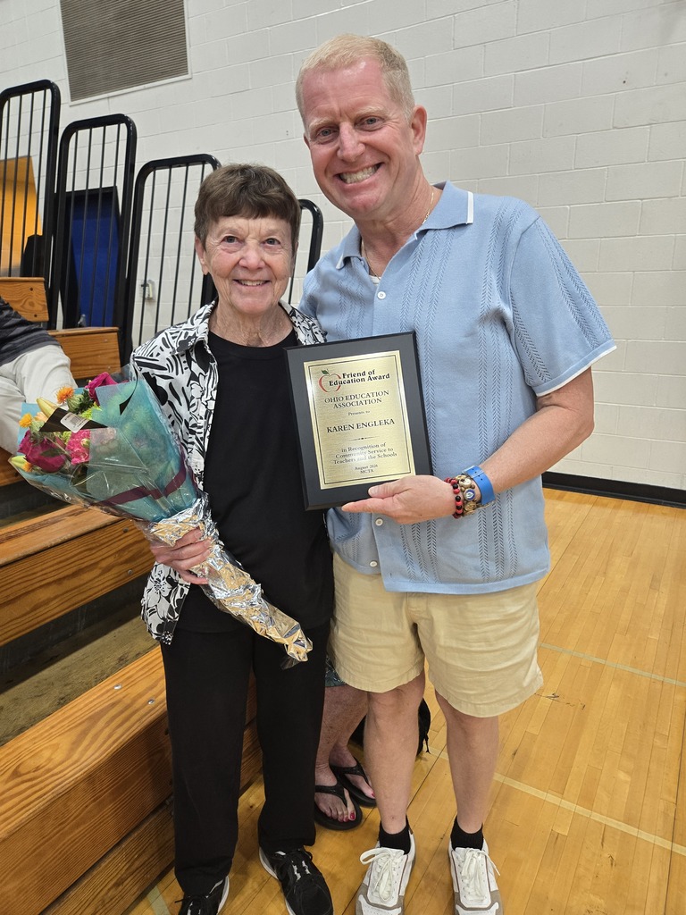 A man and woman stand, smiling, in a gymnasium. The man holds a framed plaque; the woman holds flowers. Behind them are bleachers.