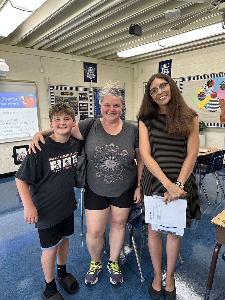 Three people stand in a classroom. A woman holds papers, a boy and a girl smile, and another woman stands with arms around them.