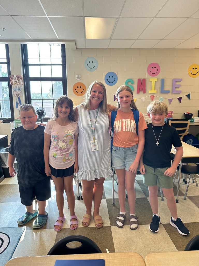 A woman with a badge stands with four children in a classroom. They stand on a checkered floor.