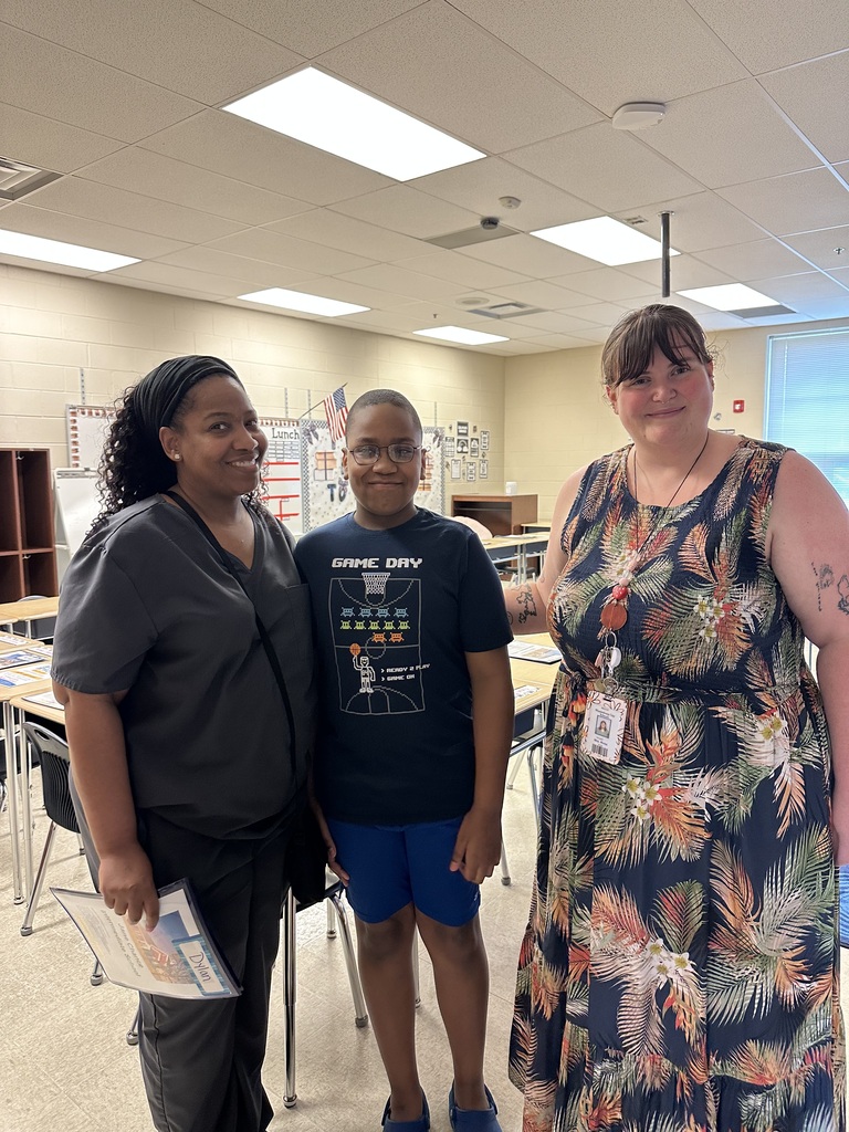 Three individuals stand in a classroom with white walls and fluorescent lighting. Two women stand on either side of a boy wearing glasses.
