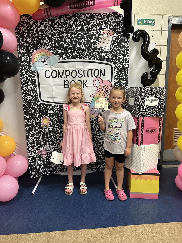 Two children stand in front of a backdrop with a composition book theme, surrounded by colorful balloons.