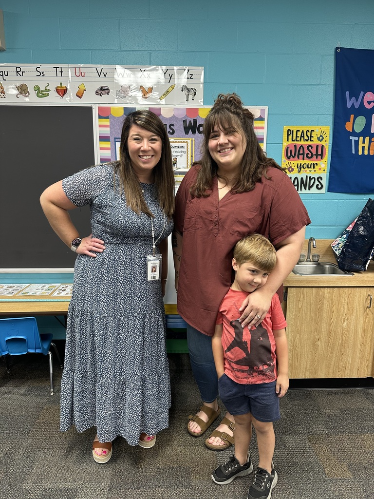 Two women and a child stand in a classroom. The woman on the left wears a dress and a watch. The other woman is in a maroon shirt. The child wears a red shirt and shorts.