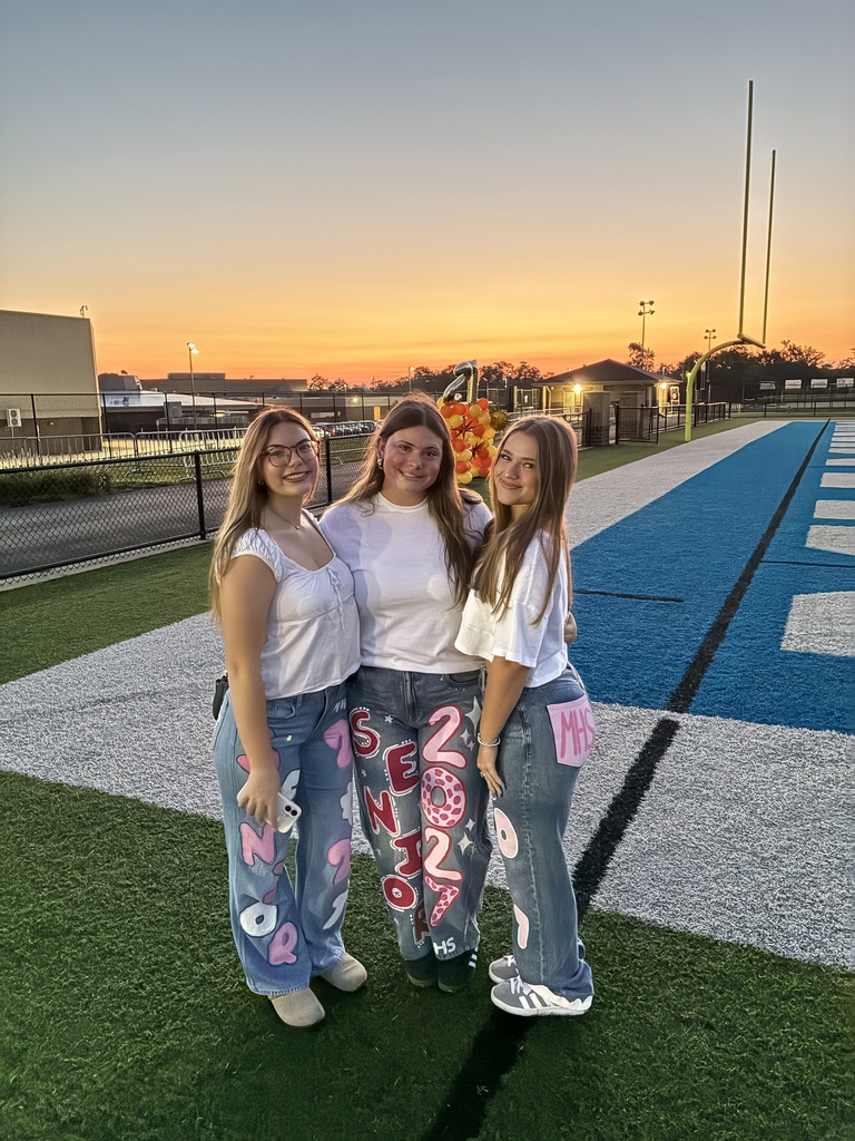 Three women in matching shirts stand on a field. One has large text on her pants. Behind them is a football field.