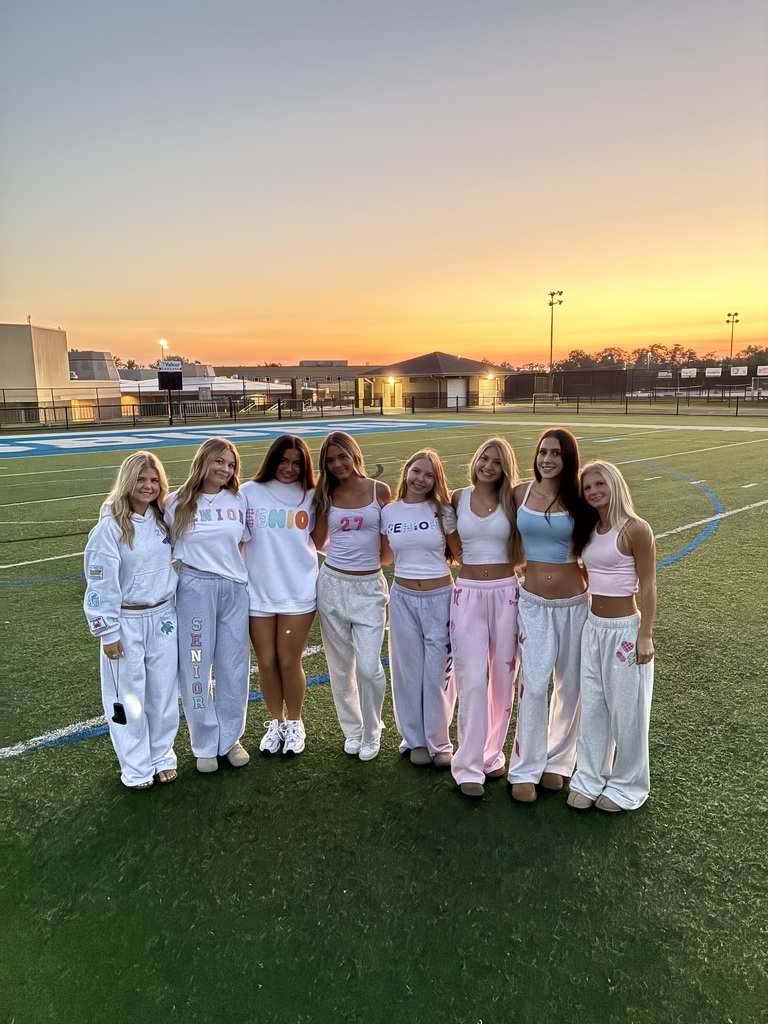 A group of women wearing matching white and pink outfits stand together on a field at sunset.