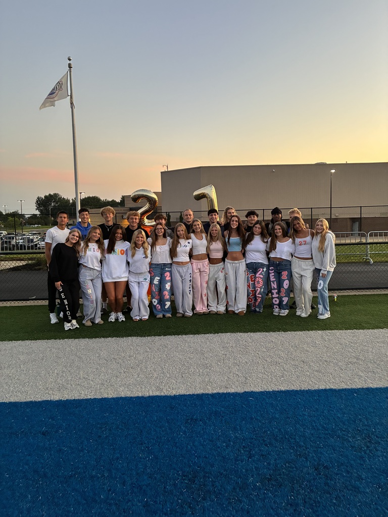 A group of people in white outfits stands on a football field with a blue and white stripe.