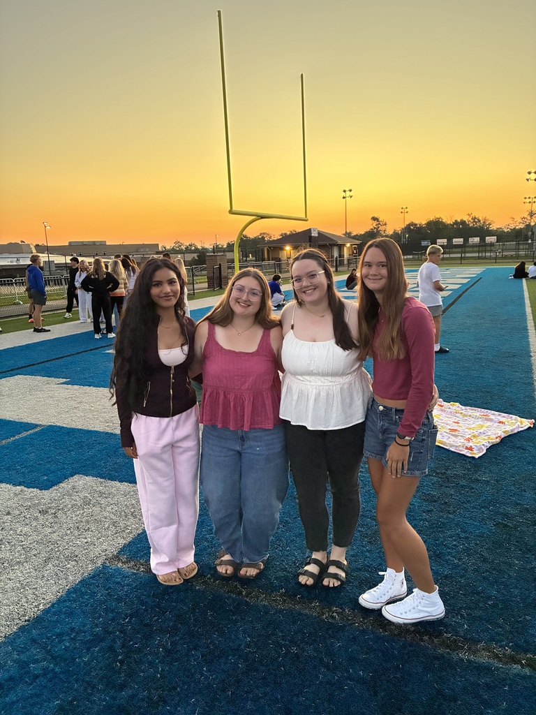 Four young women stand on a football field with a sunset in the background. They wear casual clothing.