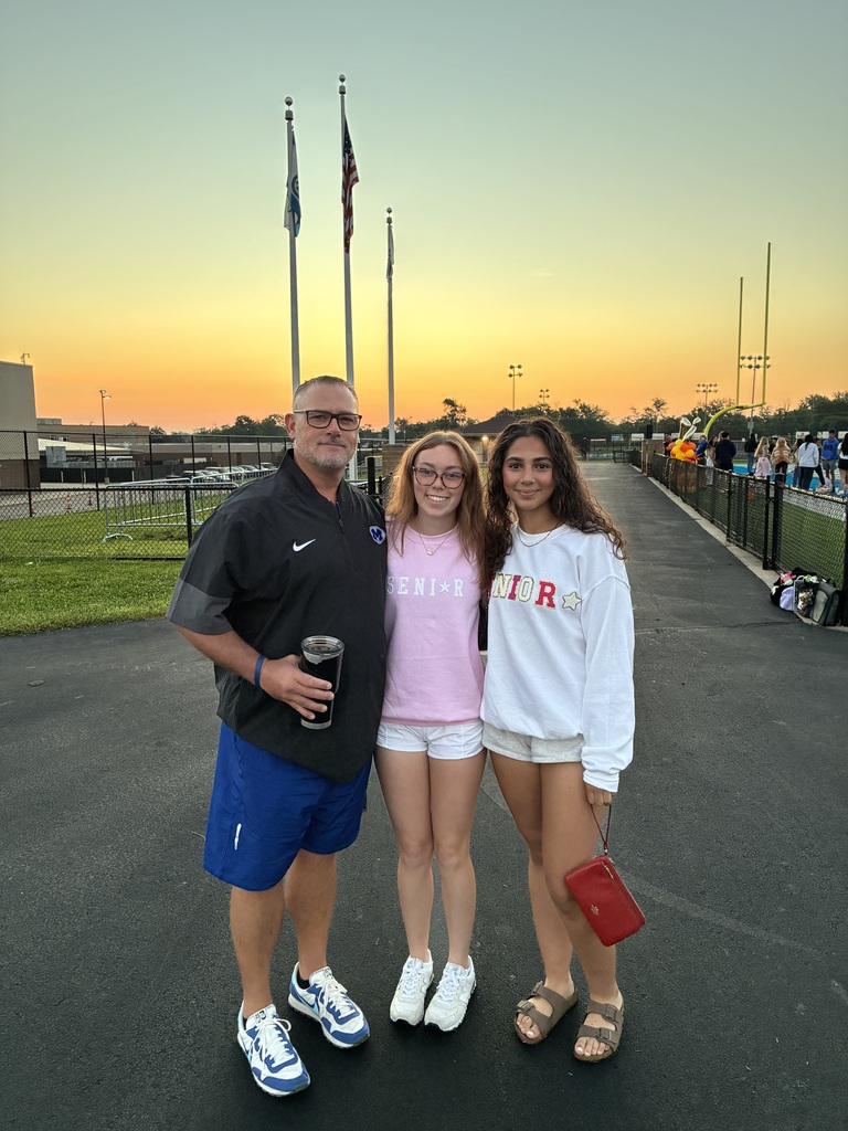 Three people stand on a road at a sports field with a sunset in the distance.