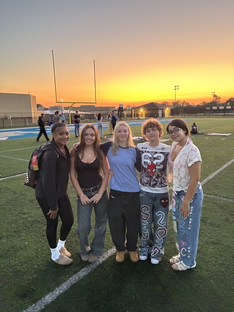 Five people stand in a line on a football field, with a sunset in the background.
