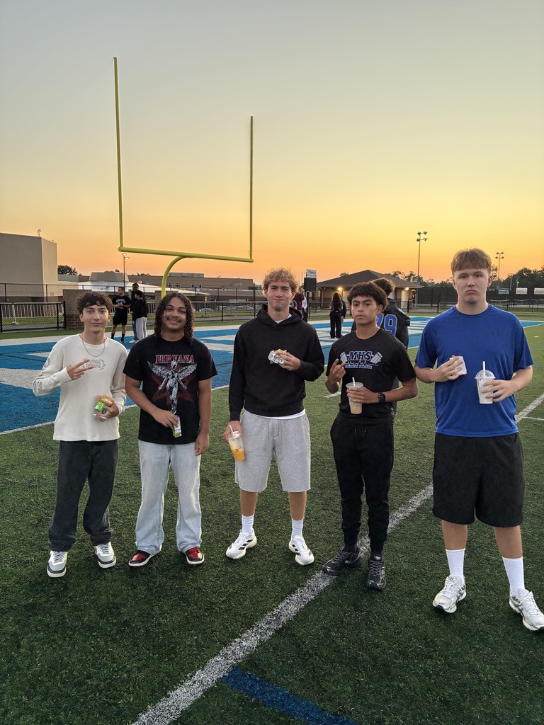Five individuals stand on a football field, holding cups and snacks, with a football goal post and sunset in the background.
