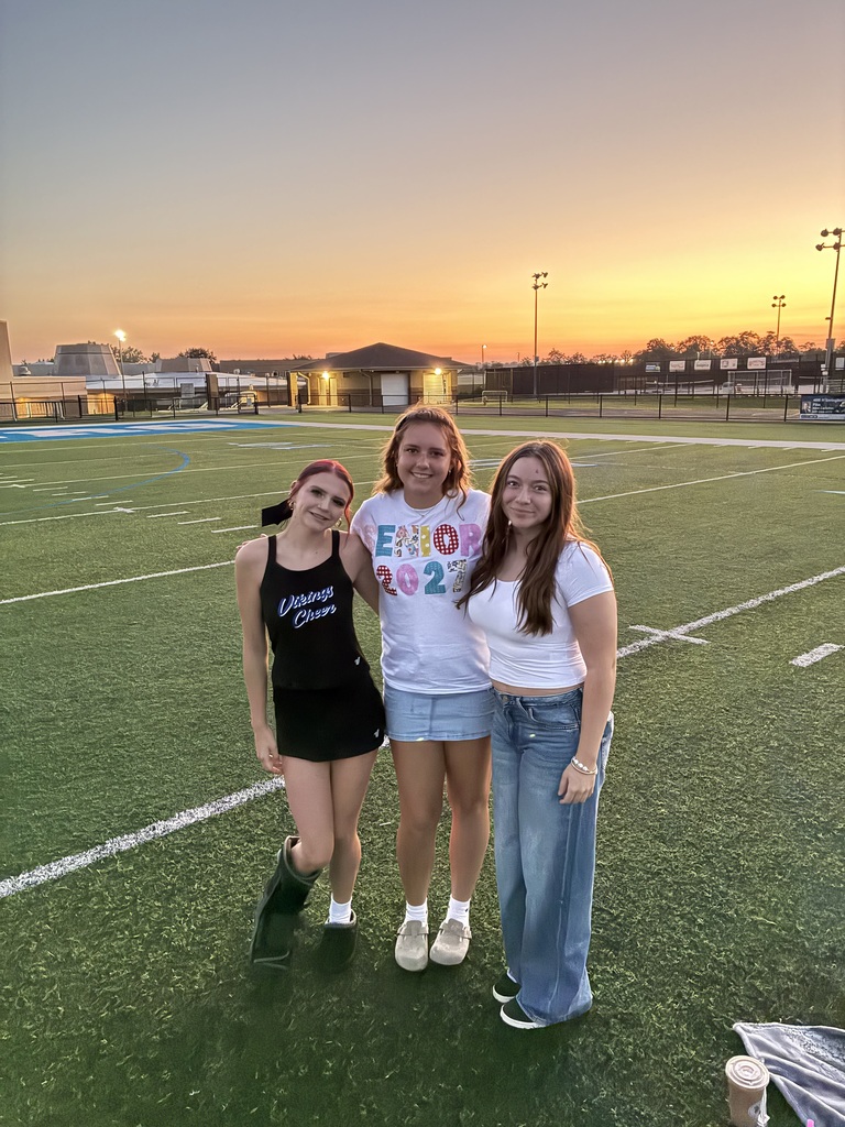 Three people pose on a field with a sunset. Two are in white tops; one wears a black dress. Field lines and distant buildings are visible.