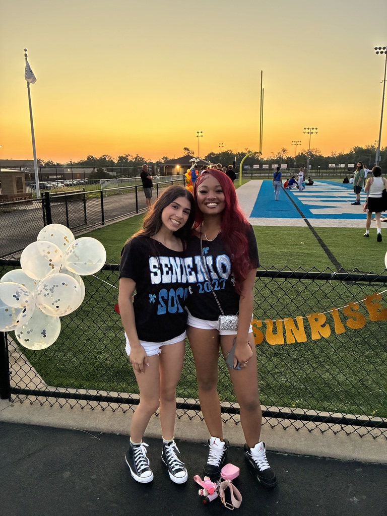 Two women stand by a fence, smiling, with balloons behind them. Behind them is a field with a sunset in the distance.