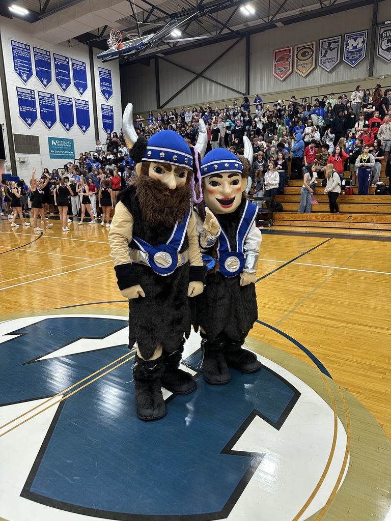 Two mascots stand on a basketball court. They wear blue and white costumes with horned helmets. An audience sits in the background.