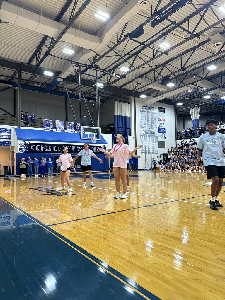A group of people play volleyball on a wooden court in a large indoor sports facility.
