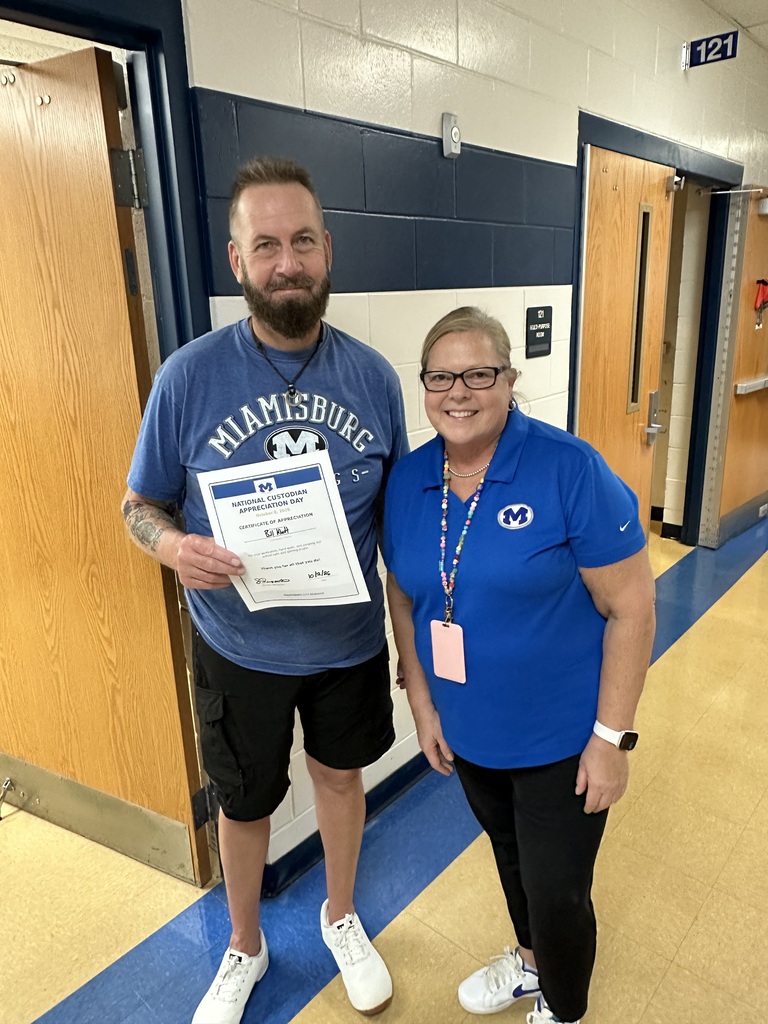 A man in a blue shirt and black shorts holds a certificate. A woman in a blue shirt and black pants stands next to him. They are in a hallway.