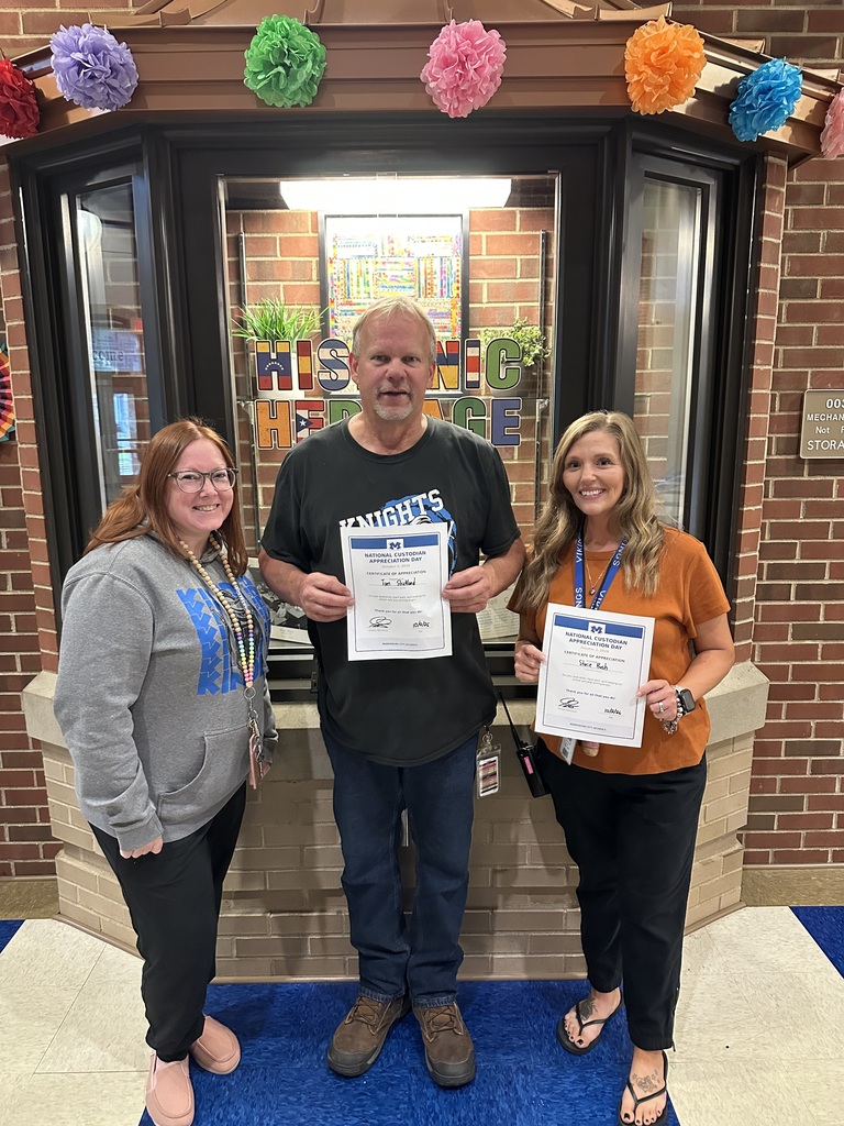 Three people stand in front of a brick building. A man holds a certificate between two women, each holding one.