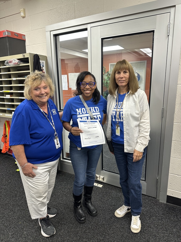 Three women stand together in front of a door. The middle woman holds a certificate. All wear blue shirts.