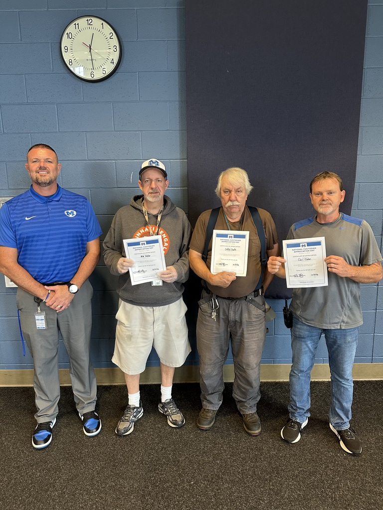 Four men stand in a room holding certificates. One wears a hat and sneakers. A clock is on a blue wall.