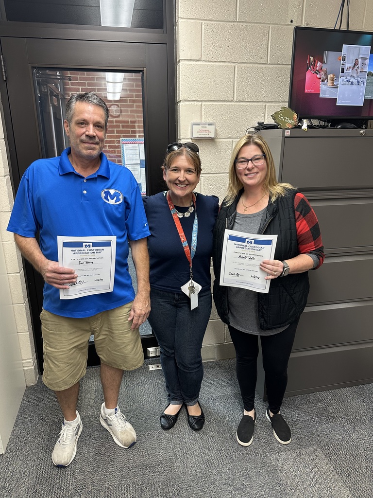 Three people stand indoors, holding certificates. One wears a blue shirt, shorts, and white sneakers. Two women wear glasses and black shoes. Behind them is a door and a TV.