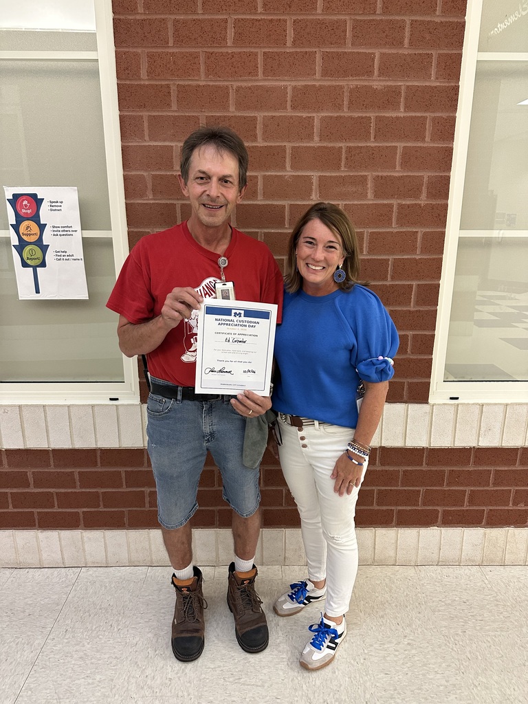 A man and a woman stand in front of a brick wall, holding a certificate. The man wears a red shirt, denim shorts, and brown boots. The woman wears a blue shirt, white pants, and blue sneakers.