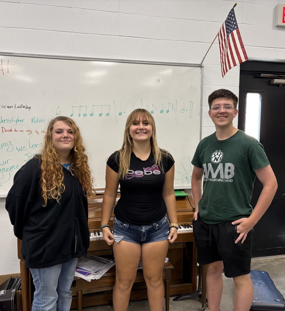 three students standing in front of a whiteboard and piano with an American flag hanging in the background