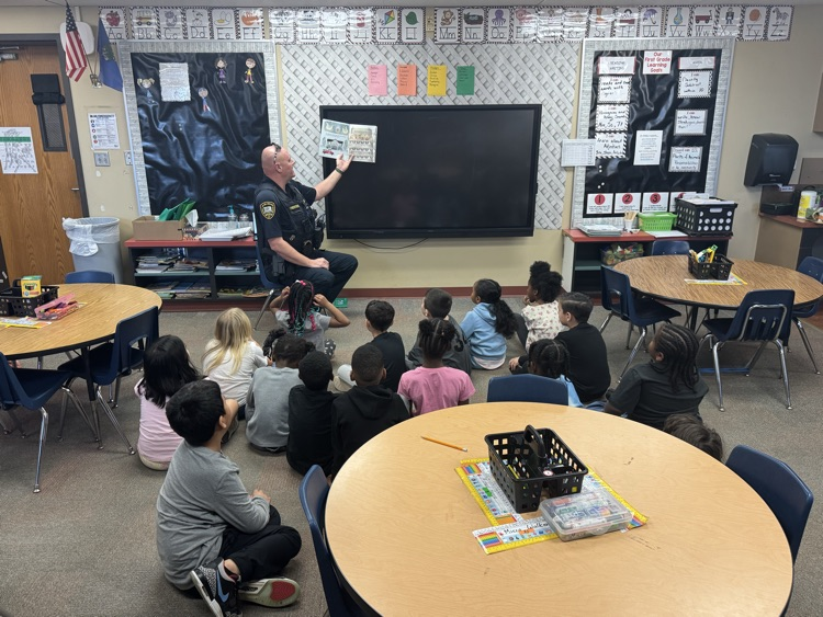 student sitting on the carpet, listening to a police officer read a book.