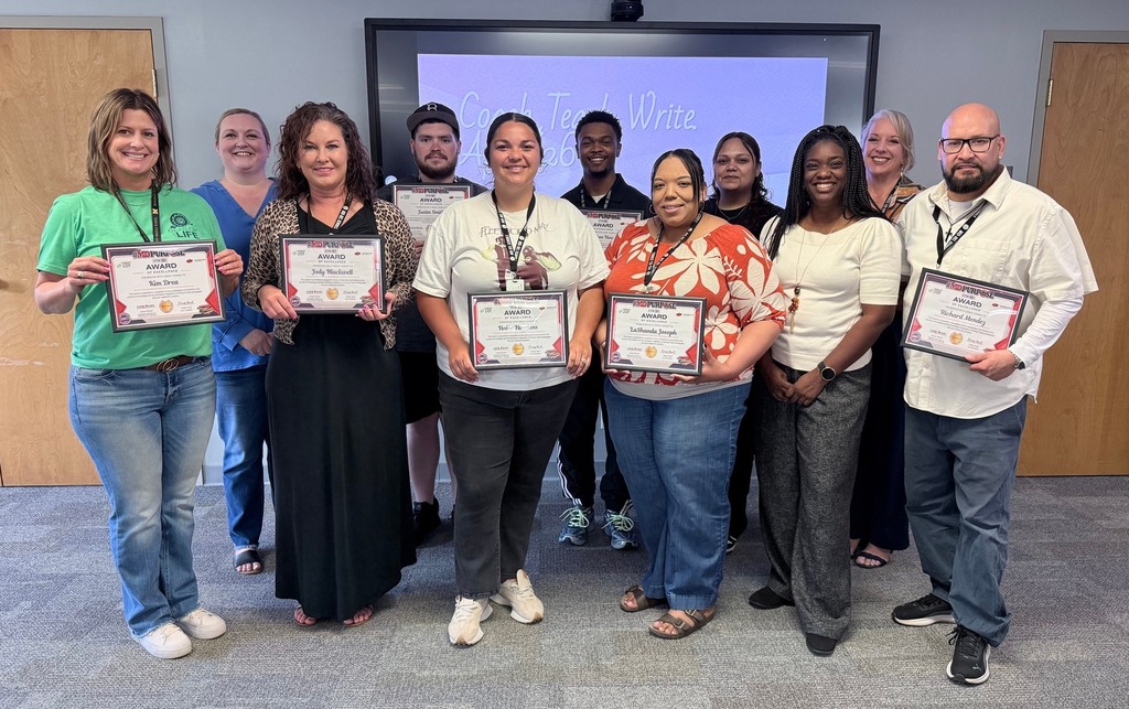 A group of ten educators from Midwest City Middle School stand together in a classroom, smiling and holding certificates of recognition. They are arranged in two rows in front of a screen displaying “Coach Teach Write,” highlighting their participation in a writing instruction grant program. The teachers are dressed in casual professional attire, and the setting appears to be a training or meeting space with doors and a presentation screen in the background.