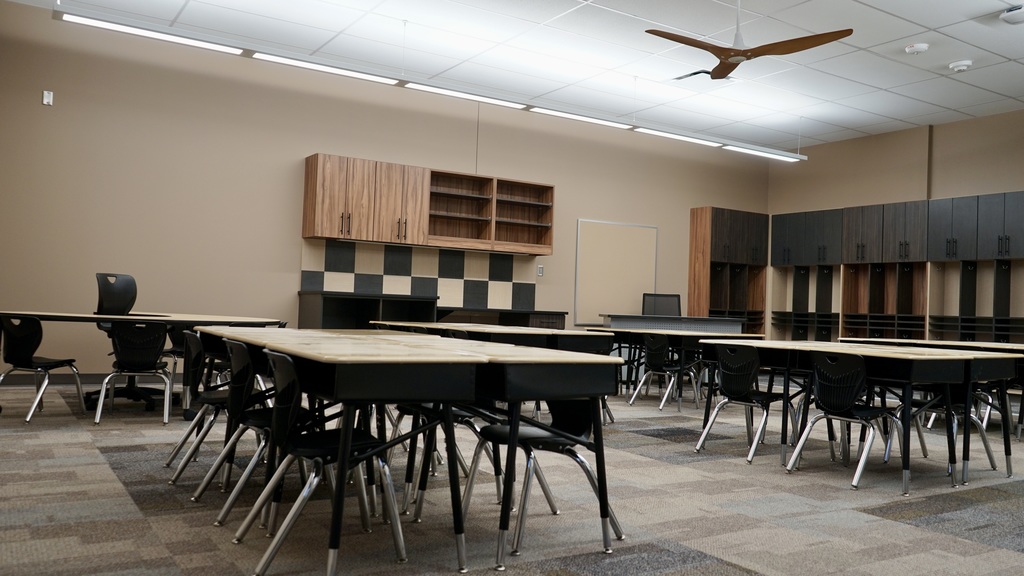 This image shows a newly finished classroom space, likely part of a modern school addition.  The room is clean, organized, and designed for collaborative learning. Several groups of student desks are arranged in clusters, each with black chairs, allowing for small-group interaction. At the front of the image, a larger teacher desk is visible.  Along the back wall, there are built-in wooden cubbies and cabinets with hooks—perfect for student storage like backpacks and jackets. There’s also a small sink area with a countertop, suggesting functionality for classroom activities. On the right wall, additional cabinetry and shelving provide extra storage, along with a whiteboard for instruction.  The flooring is a mix of patterned carpet tiles, adding a modern, durable touch. Overall, the space feels bright, updated, and thoughtfully designed to support both learning and organization.