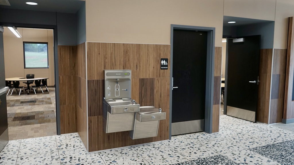 This image shows a newly finished classroom space, likely part of a modern school addition.  The room is clean, organized, and designed for collaborative learning. Several groups of student desks are arranged in clusters, each with black chairs, allowing for small-group interaction. At the front of the image, a larger teacher desk is visible.  Along the back wall, there are built-in wooden cubbies and cabinets with hooks—perfect for student storage like backpacks and jackets. There’s also a small sink area with a countertop, suggesting functionality for classroom activities. On the right wall, additional cabinetry and shelving provide extra storage, along with a whiteboard for instruction.  The flooring is a mix of patterned carpet tiles, adding a modern, durable touch. Overall, the space feels bright, updated, and thoughtfully designed to support both learning and organization.