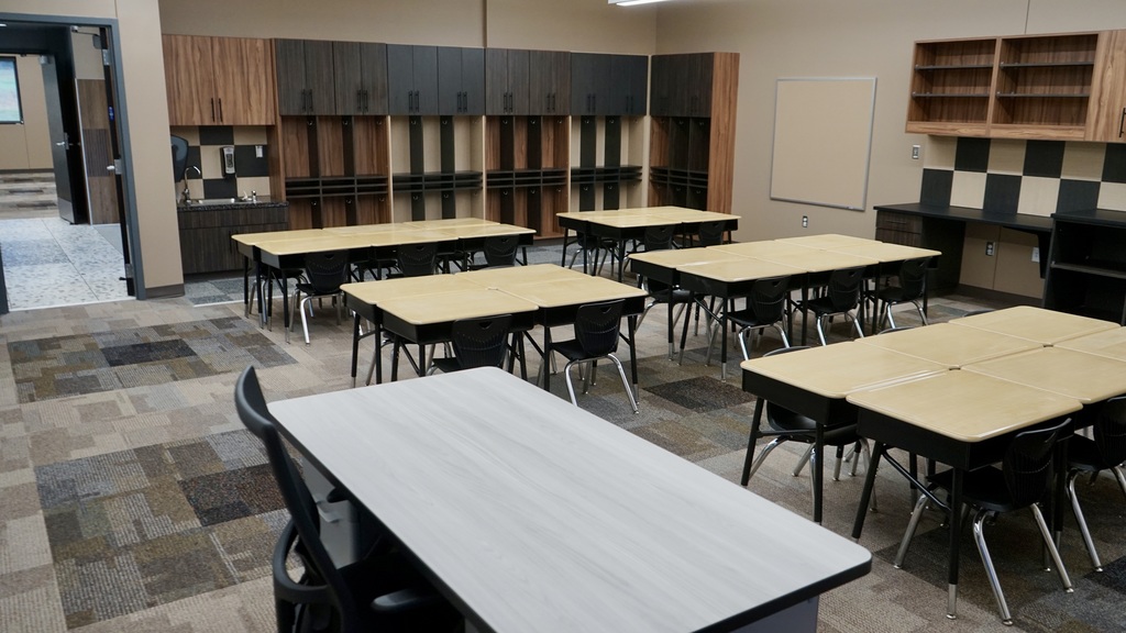 This image shows a newly finished classroom space, likely part of a modern school addition.  The room is clean, organized, and designed for collaborative learning. Several groups of student desks are arranged in clusters, each with black chairs, allowing for small-group interaction. At the front of the image, a larger teacher desk is visible.  Along the back wall, there are built-in wooden cubbies and cabinets with hooks—perfect for student storage like backpacks and jackets. There’s also a small sink area with a countertop, suggesting functionality for classroom activities. On the right wall, additional cabinetry and shelving provide extra storage, along with a whiteboard for instruction.  The flooring is a mix of patterned carpet tiles, adding a modern, durable touch. Overall, the space feels bright, updated, and thoughtfully designed to support both learning and organization.
