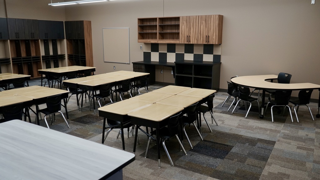 This image shows a newly finished classroom space, likely part of a modern school addition.  The room is clean, organized, and designed for collaborative learning. Several groups of student desks are arranged in clusters, each with black chairs, allowing for small-group interaction. At the front of the image, a larger teacher desk is visible.  Along the back wall, there are built-in wooden cubbies and cabinets with hooks—perfect for student storage like backpacks and jackets. There’s also a small sink area with a countertop, suggesting functionality for classroom activities. On the right wall, additional cabinetry and shelving provide extra storage, along with a whiteboard for instruction.  The flooring is a mix of patterned carpet tiles, adding a modern, durable touch. Overall, the space feels bright, updated, and thoughtfully designed to support both learning and organization.