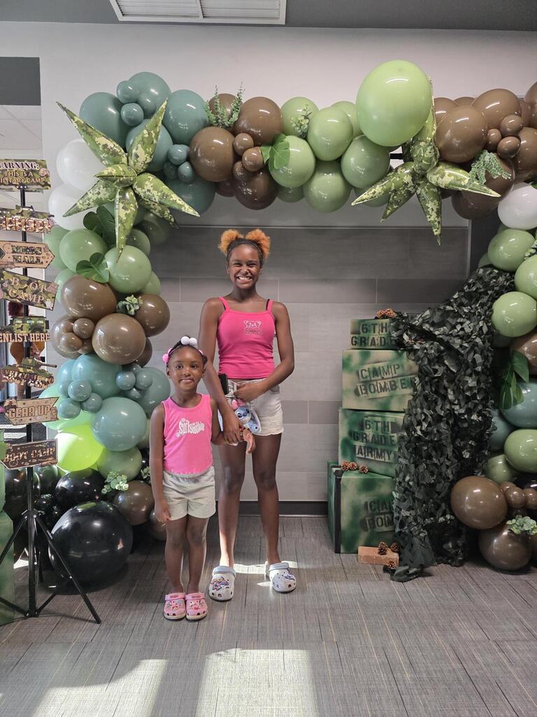 a photo of 2 young ladies standing in front of a camo baloon arch