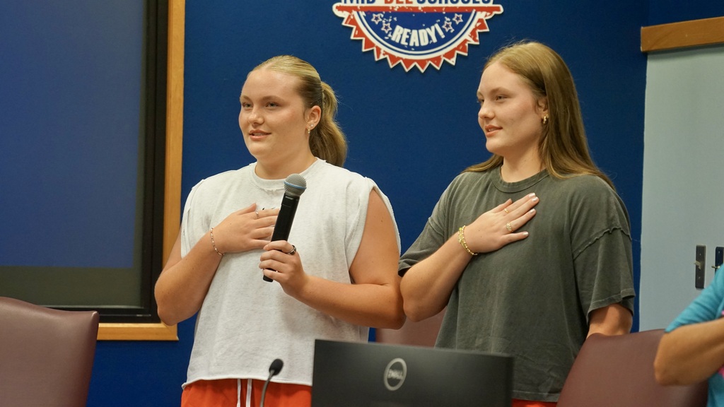 Two women standing, one holding a microphone, and the other placing her hand on her chest.