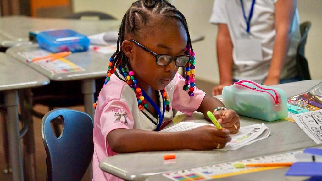 A young girl in glasses sits at a desk, holding a pen and coloring a paper.