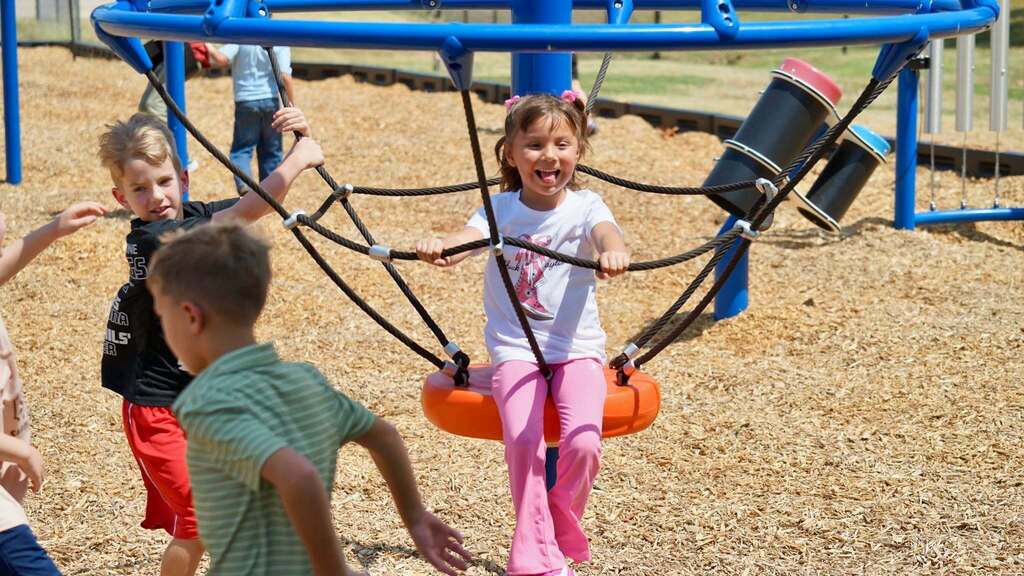 Kids playing on a swing set. One child holds onto the ropes, another swings on the swing, and others run nearby.