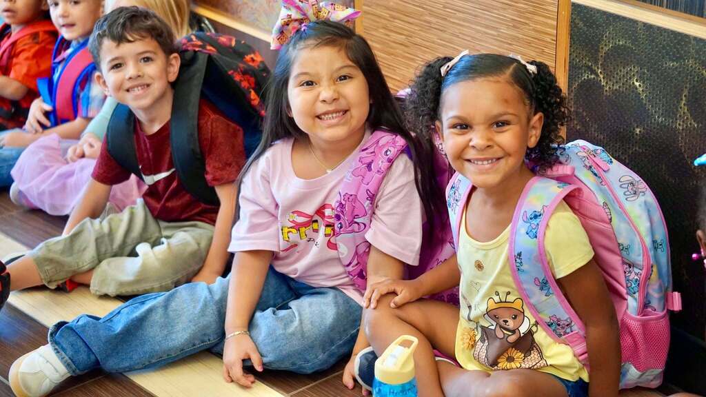 Four children, wearing backpacks, seated on the floor in a classroom. Two girls smile and pose.