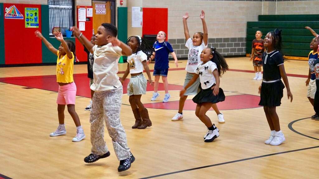 Several children dance on a basketball court, with two in raised hands. Some wear sneakers; others have skirts. The floor is wooden with red markings.