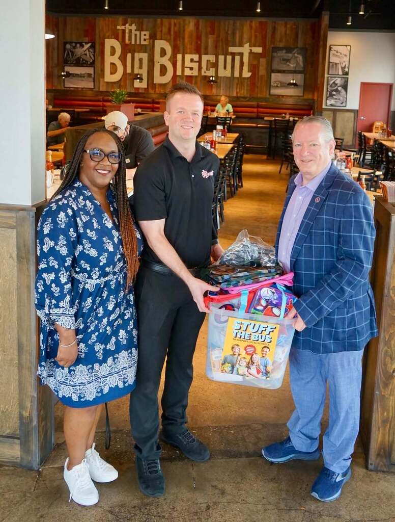 Three people stand in a restaurant with a banner reading "The Big Biscuit". They hold a toy box.