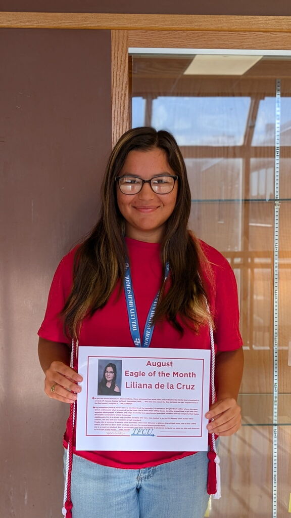 Woman standing in front of a display case holding a certificate, reading "August Eagle of the Month Liliana de la Cruz."